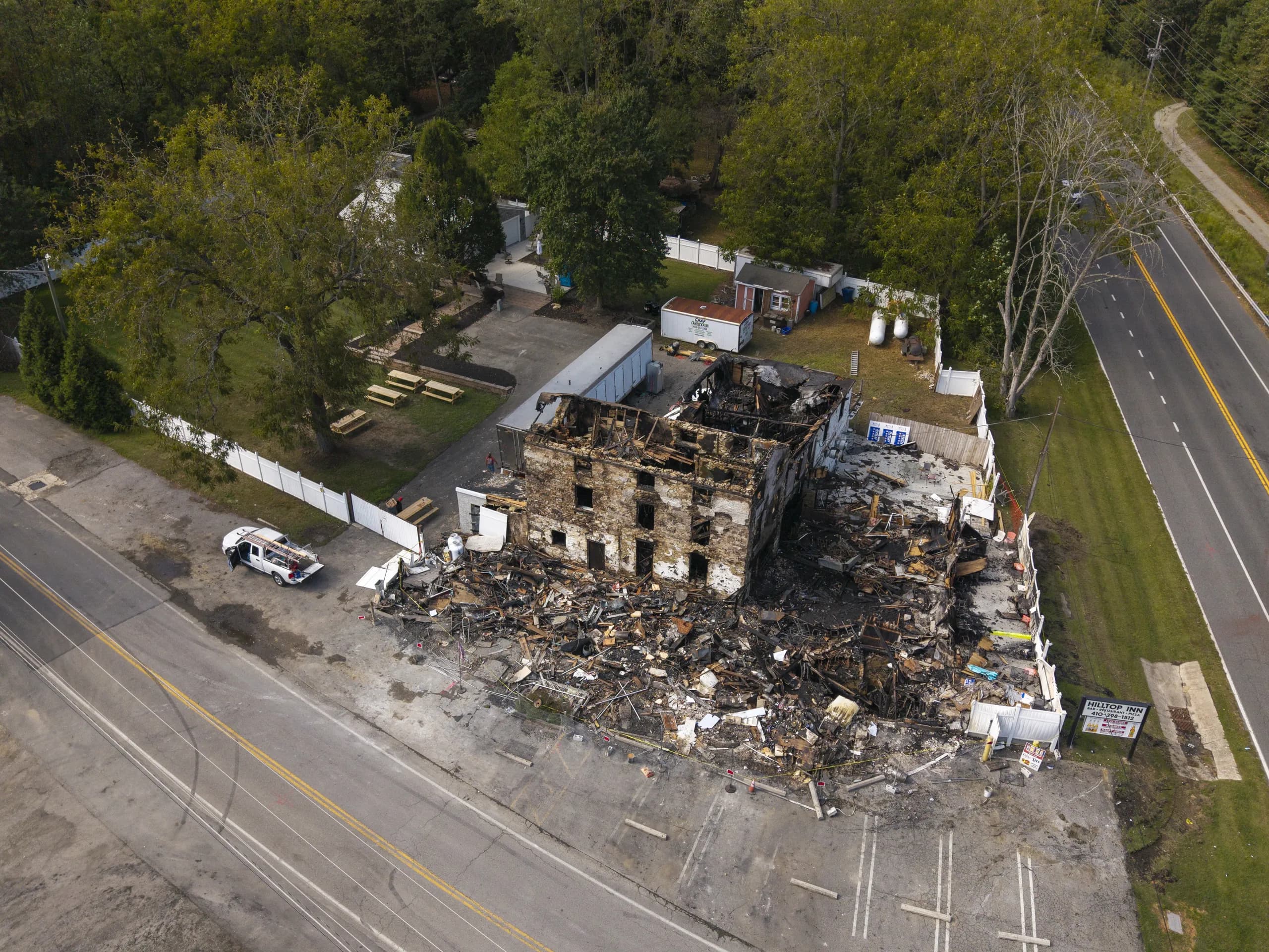 Aerial oblique of a burnt-out two-storey roadside inn, the masonry walls standing above a debris field of charred timber, with the road and parking area in frame